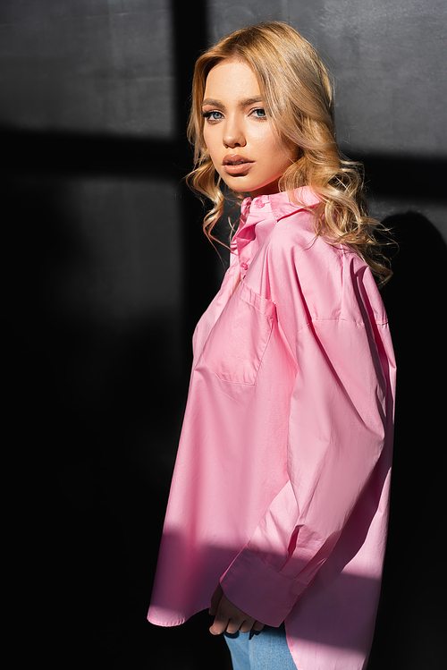 young woman in pink shirt looking at camera near black wall with shadows