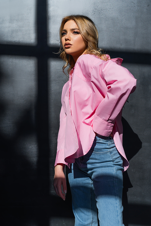 stylish woman in pink shirt standing with hand in pocket of jeans near black wall with shadows