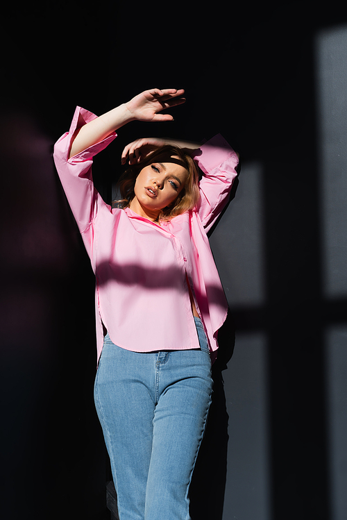 young and stylish woman standing with hands above head on black background with shadows