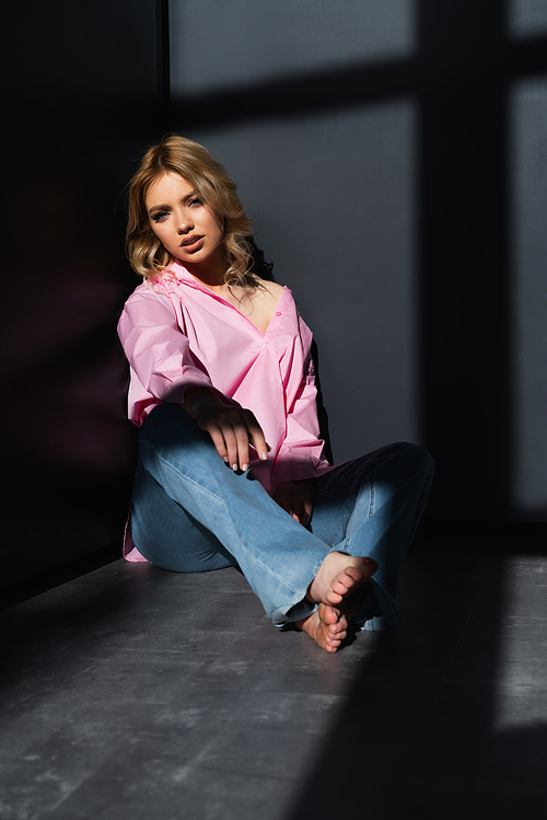 full length view of barefoot woman in jeans and pink shirt sitting in corner near black walls