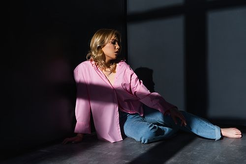 full length view of barefoot woman in jeans sitting in lighting between black walls
