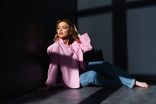 full length of barefoot woman in pink shirt sitting on floor near black walls with shadows