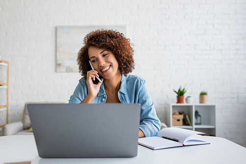 Positive african american freelancer talking on cellphone and looking at laptop in living room