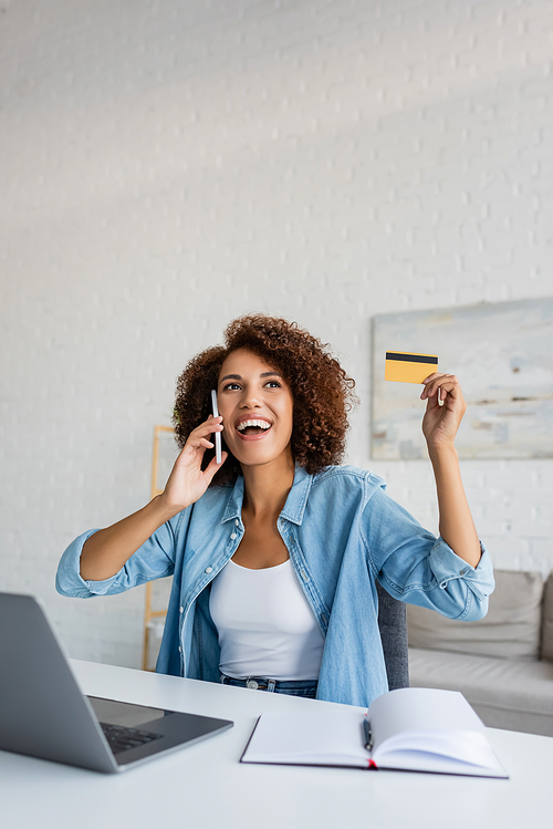 Young african american woman holding credit card and talking on smartphone at home