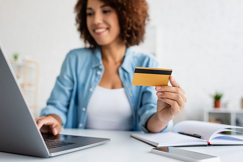 Blurred african american woman holding credit card and using laptop near notebook on table