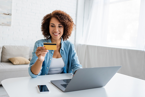 Positive african american woman holding credit card near devices at home