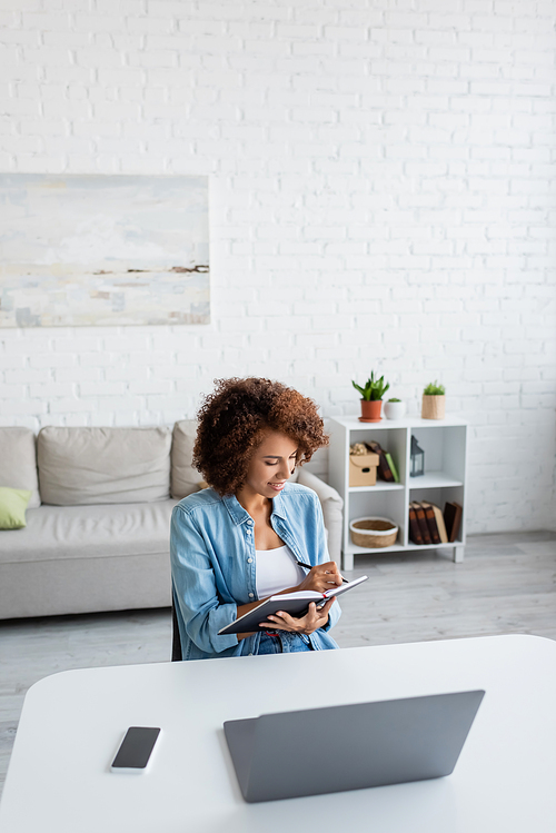 Curly african american freelancer writing on notebook near devices in living room