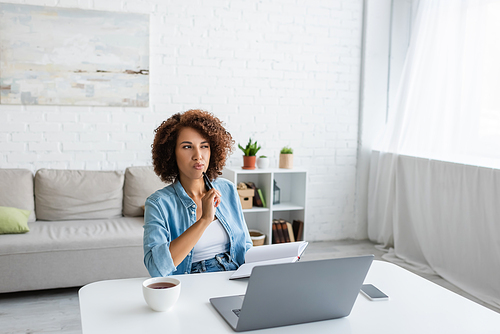 Pensive african american freelancer holding notebook near gadgets and tea in living room