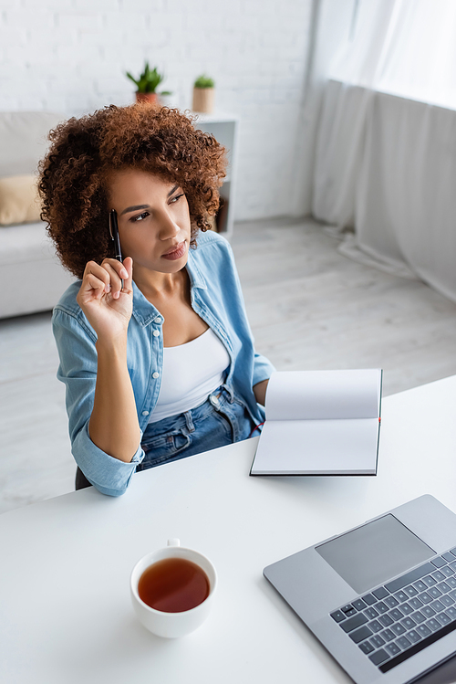 Thoughtful african american freelancer holding notebook near laptop and cup of tea at home