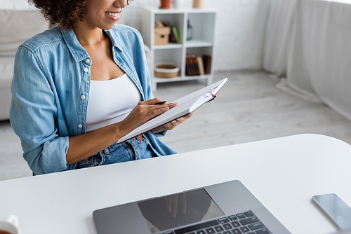 Cropped view of cheerful african american freelancer holding notebook near devices with blank screen at home