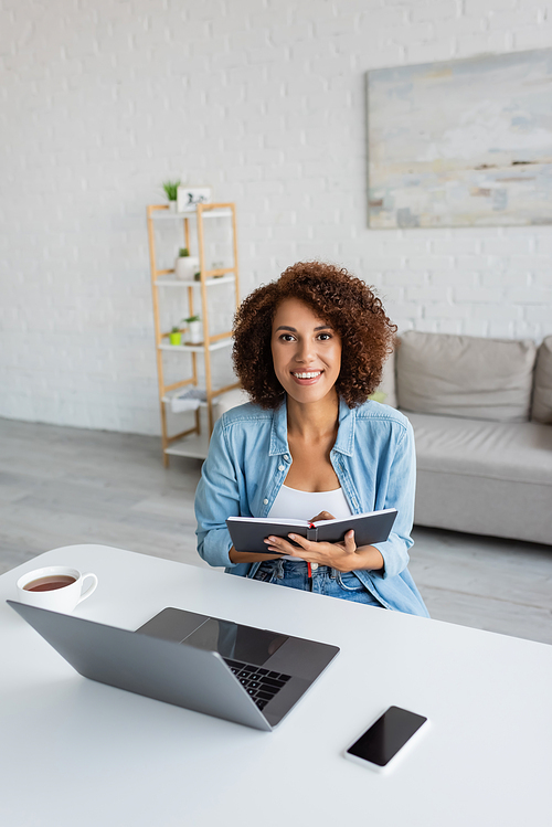 Positive african american woman looking at camera near holding notebook near devices and tea at home