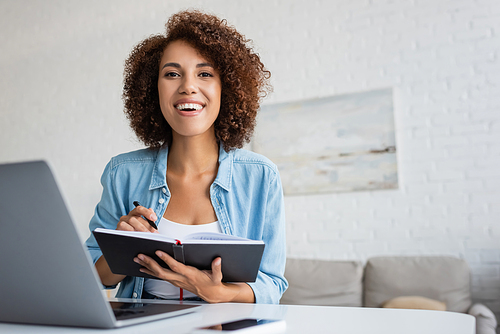 Happy african american freelancer looking at camera and holding notebook at home