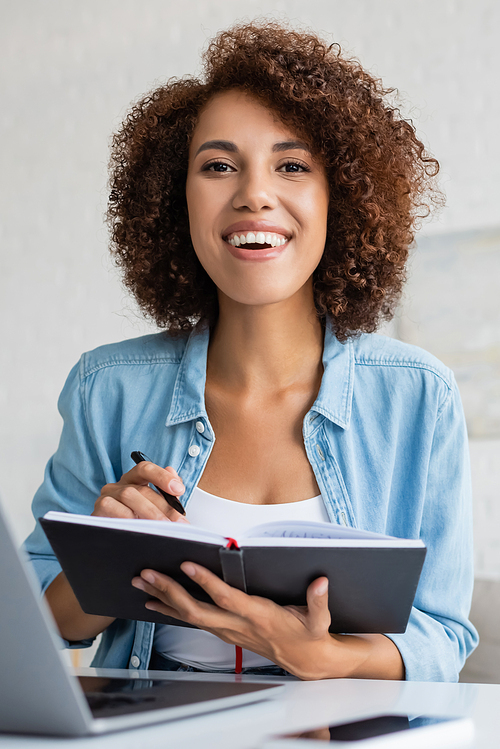 Portrait of positive african american freelancer holding notebook near blurred gadgets at home