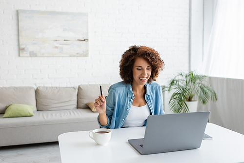 Smiling african american freelancer holding pen near devices and cup of tea at home