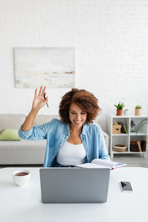 Smiling african american freelancer with notebook waving hand during video call on laptop at home