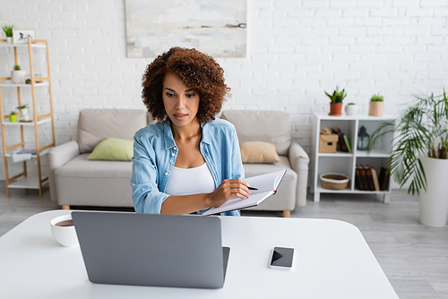 Curly african american woman holding notebook and using laptop near cup of tea at home