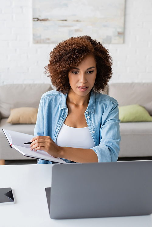 Young african american freelancer holding notebook while using laptop near smartphone on table at home