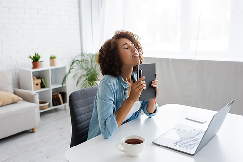 Stressed african american freelancer holding notebook near devices and tea at home