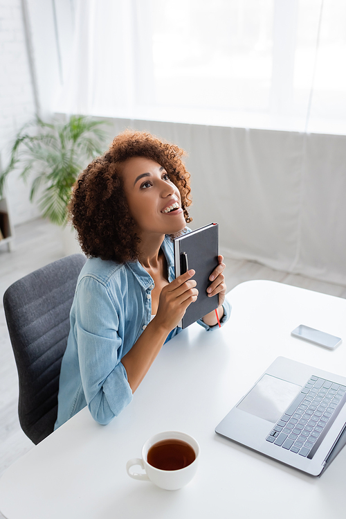 Positive african american woman holding notebook and looking up near devices and tea in living room