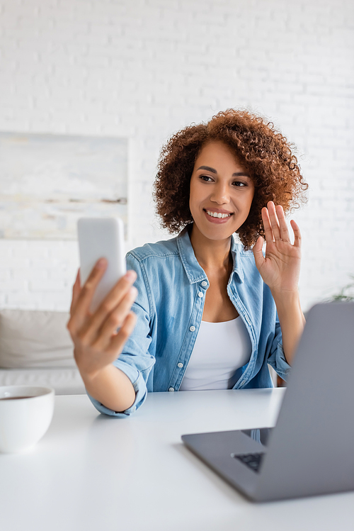 Curly african american woman having video call on smartphone near laptop and tea at home