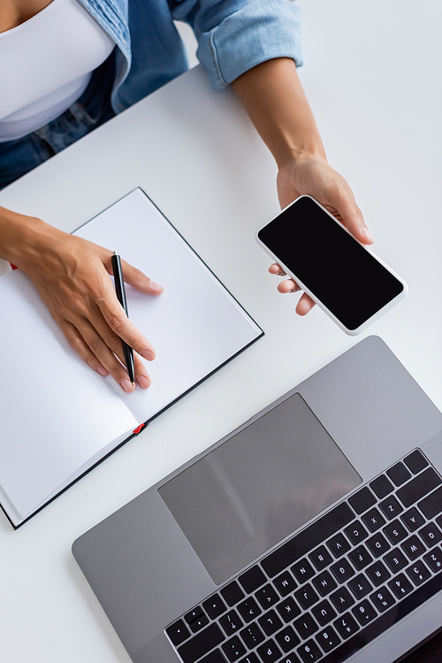 Top view of african american woman holding smartphone with blank screen near laptop and notebook at home