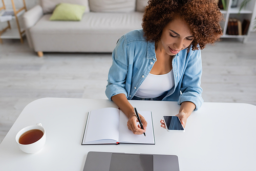 African american freelancer using mobile phone and writing on notebook near tea at home