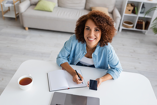 Cheerful african american woman holding smartphone and writing on notebook near laptop at home