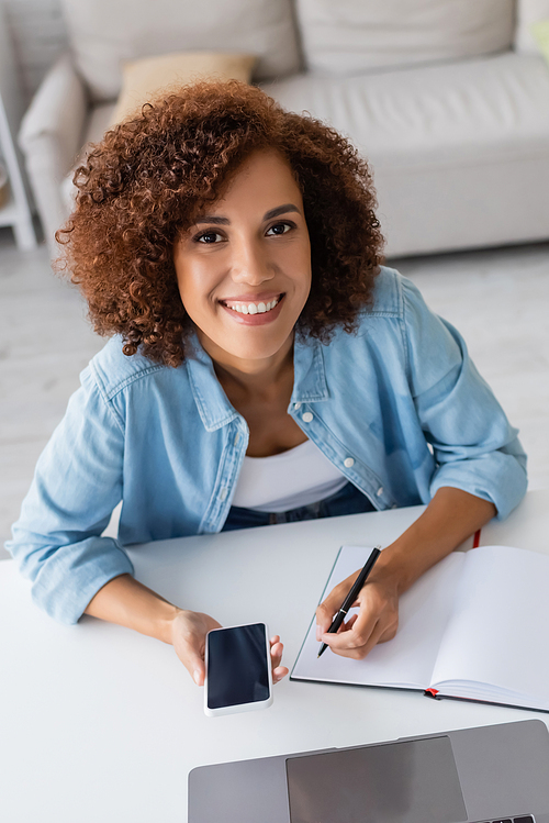 High angle view of african american freelancer holding smartphone near notebook at home