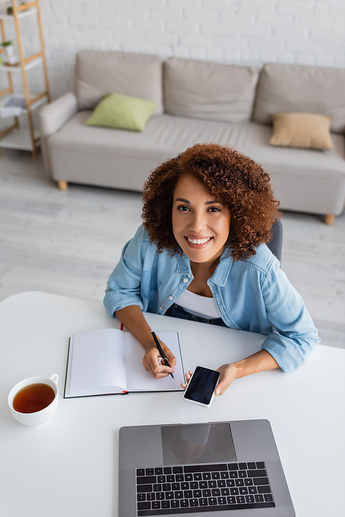 High angle view of positive freelancer using devices and writing on notebook near cup of tea at home