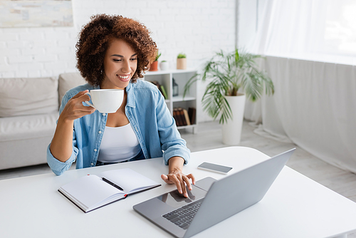 Positive african american woman holding cup and using laptop near notebook on table at home