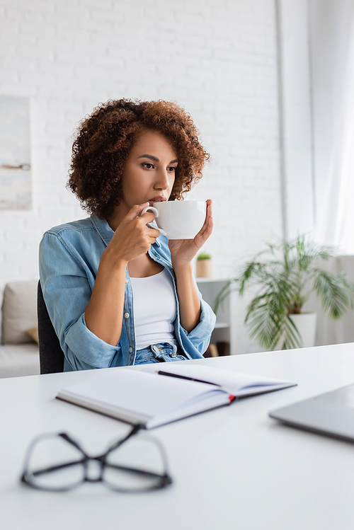 curly african american woman drinking coffee and looking at laptop on desk