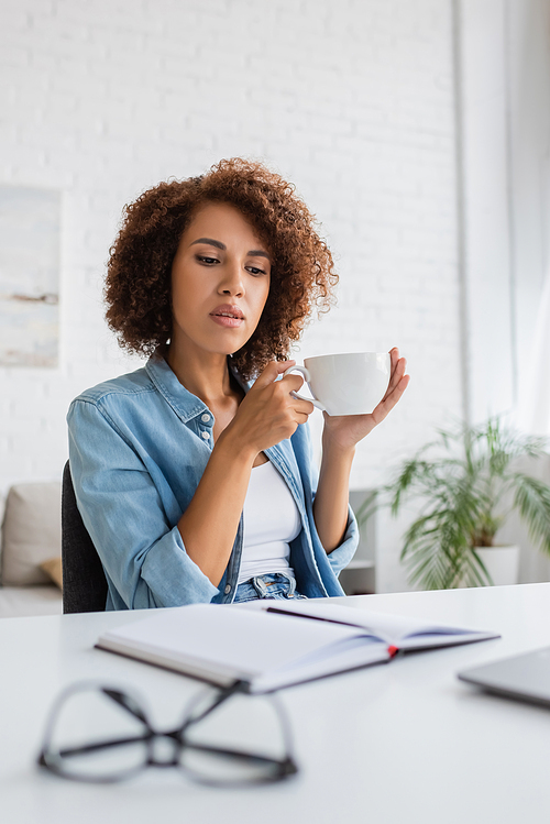 pensive african american woman holding cup of coffee and looking at notebook on desk