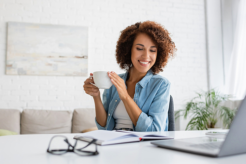 cheerful african american woman holding cup of coffee and looking at notebook near laptop on desk