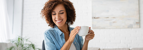 cheerful african american woman holding cup of coffee and looking away at home, banner