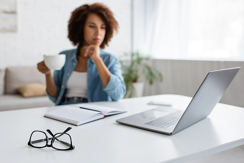 laptop and stationery on desk near blurred african american freelancer with cup