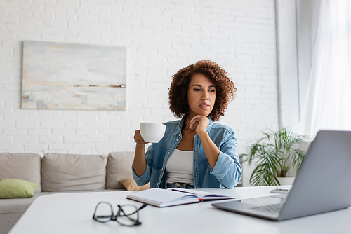 pensive african american woman holding cup of coffee and looking at laptop on desk