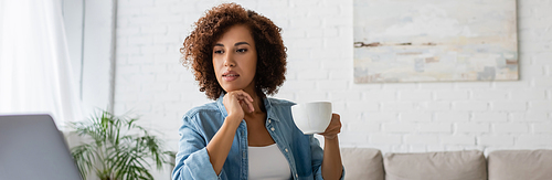 pensive african american woman holding cup of coffee and looking at laptop, banner