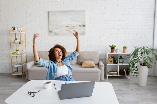 pleased african american woman with closed eyes rejoicing near workplace at home