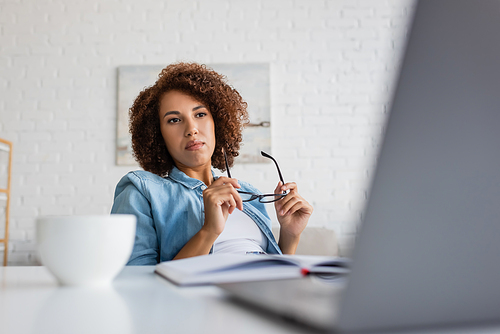 curly african american woman holding glasses and looking at blurred laptop