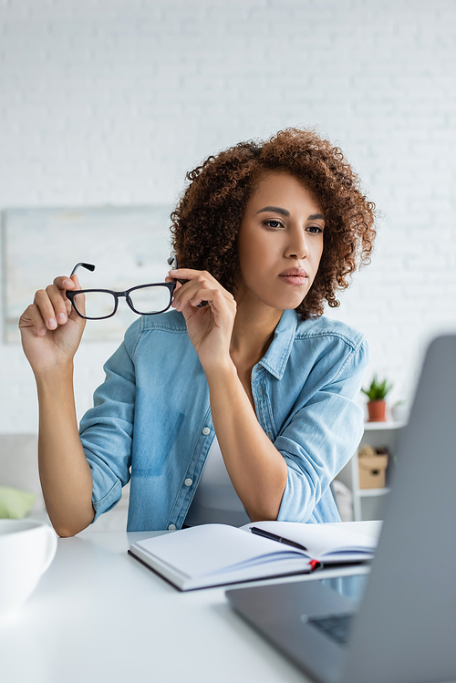 curly african american woman holding glasses and looking at blurred laptop on table