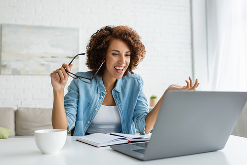 happy african american woman holding glasses and looking at laptop on table