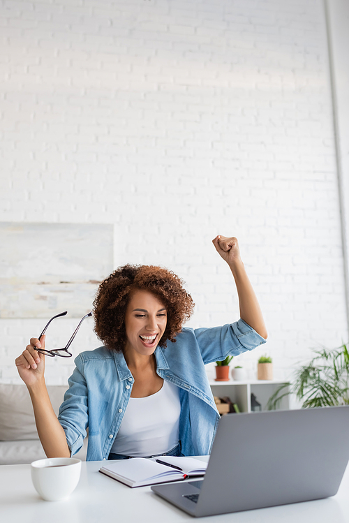 excited african american woman holding glasses and looking at laptop on table