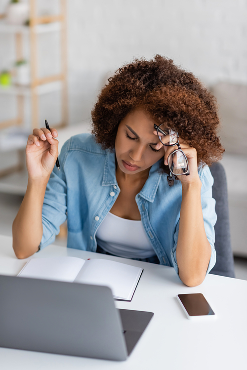 tired african american woman holding glasses and pen near gadgets on desk