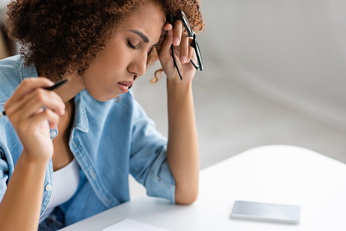 tired african american woman holding glasses and pen near smartphone with blank screen on desk