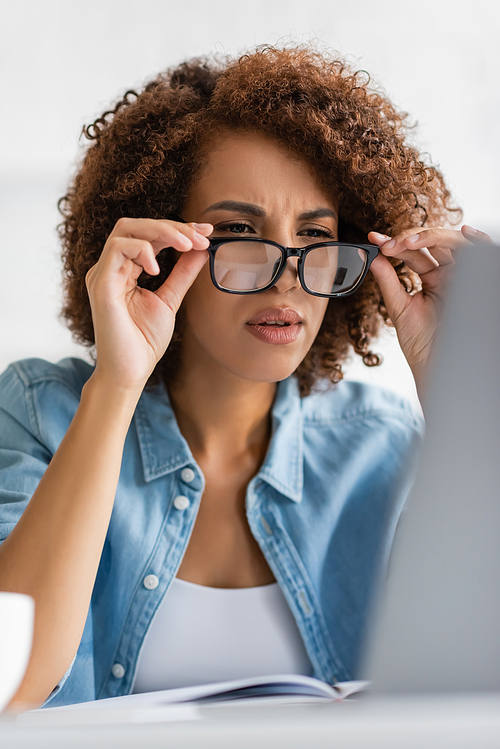 focused african american woman wearing glasses and looking at blurred laptop