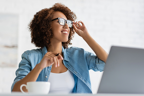 happy african american woman wearing glasses and looking away near laptop