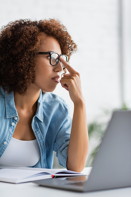 curly african american woman adjusting glasses and looking away near blurred laptop