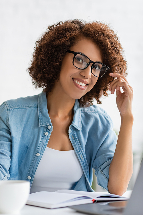 happy african american woman in glasses looking at camera while sitting at desk