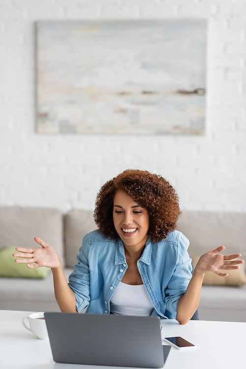 cheerful and curly african american freelancer gesturing during video call on laptop