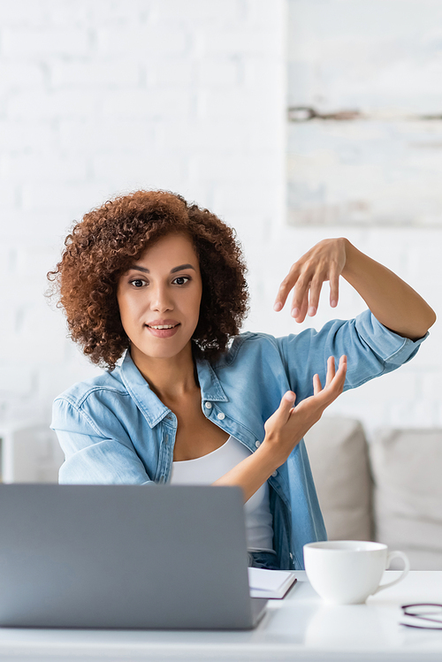 curly african american woman gesturing during video call on laptop near cup on desk